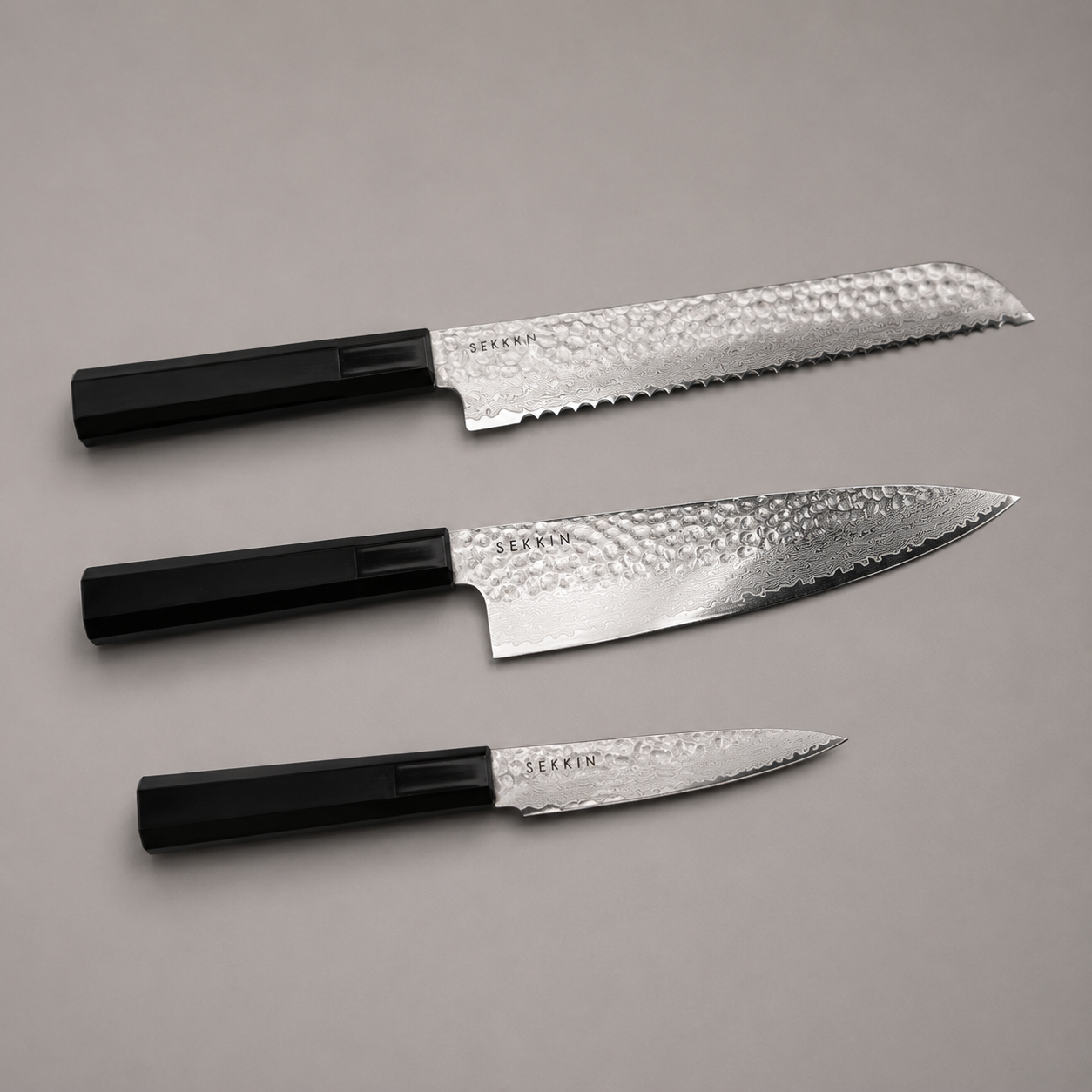 Three kitchen knives with textured silver blades and black handles, arranged diagonally on a neutral gray background.